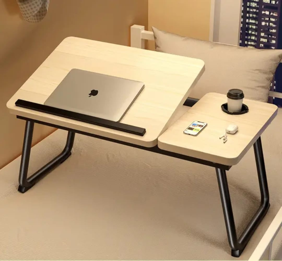 A light wood and black metal lap desk shown on a bed, with a laptop on the main desk surface and a small side tray holding a cup, phone, earbuds case, and keys.