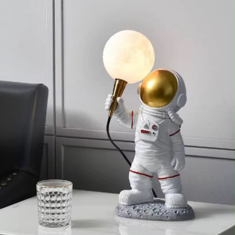 A novelty astronaut-shaped table lamp with a glowing spherical moon globe and a metallic gold helmet, displayed on a white tabletop next to a textured glass.